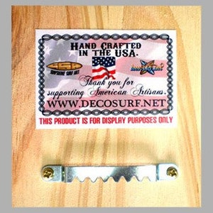 Op de afbeelding: Achterkant van een houten bord met een metalen ophanghaak. Het bord heeft een wit label met de tekst "HAND CRAFTED IN THE USA. Thank you for supporting American Artisans. WWW.DECOSURF.NET THIS PRODUCT IS FOR DISPLAY PURPOSES ONLY".