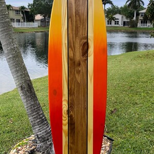 May include: A wooden surfboard painted with a gradient of orange, yellow, and red. The surfboard has three black stripes down the center.