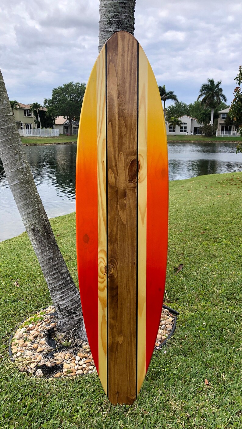 May include: A wooden surfboard painted with a gradient of orange, yellow, and red. The surfboard has three black stripes down the center.
