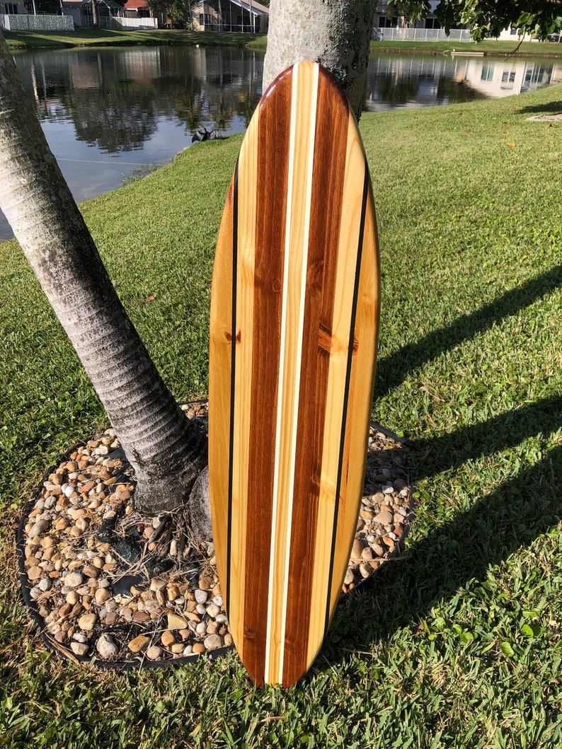 Op de afbeelding: Een houten surfboard met een bruin, wit en zwart gestreept ontwerp. Het surfboard leunt tegen een boomstam.