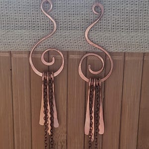 Op de afbeelding: Koperkleurige oorhangers. Elke oorbel heeft een gebogen bovenkant met een spiraalvormig ontwerp, verbonden met een reeks kleine kettingen en gehamerde metalen vormen. De oorbellen worden gepresenteerd tegen een bamboe achtergrond.