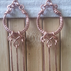 May include: A pair of handcrafted copper earrings. Each earring features a woven copper hoop with a decorative knot and three dangling copper bars. The earrings are displayed against a light gray fabric and a wooden background.