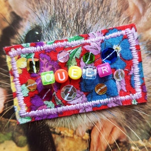 May include: A colourful fabric patch with a floral pattern and the word "QUEER" spelled out in rainbow letter beads.