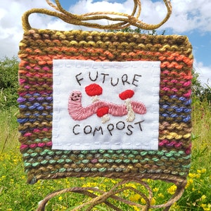 以下が含まれることがあります： 白い布のパネルに「FUTURE COMPOST」の文字と、赤いキノコをつけたピンクのミミズが刺繍された、織りの壁掛け。織りの背景は、茶色、緑、オレンジ、ピンク、紫が混ざり合っています。