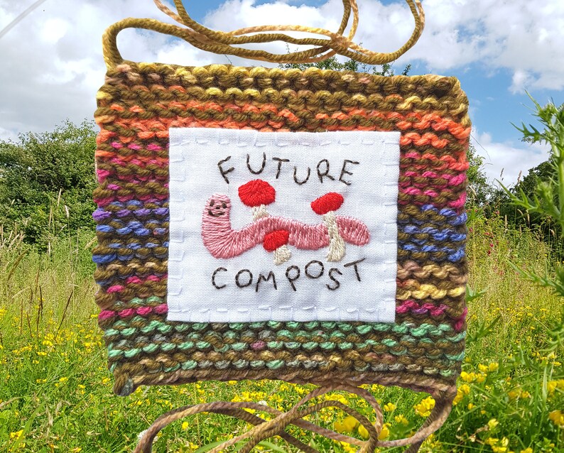 以下が含まれることがあります： 白い布のパネルに「FUTURE COMPOST」の文字と、赤いキノコをつけたピンクのミミズが刺繍された、織りの壁掛け。織りの背景は、茶色、緑、オレンジ、ピンク、紫が混ざり合っています。