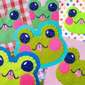 May include: A collection of six felt frog patches with embroidered details. The frogs have green bodies, pink cheeks, blue eyes, and pink tongues. The patches are sewn onto different fabric backgrounds.