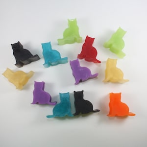 Op de afbeelding: Een verzameling van 12 kleine, kleurrijke kattenbeeldjes. De beeldjes zijn gemaakt van een glad, glanzend materiaal en zijn in een cirkel op een wit oppervlak gerangschikt. De kleuren omvatten rood, oranje, geel, groen, blauw, paars en zwart.