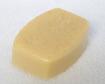 May include: A rectangular bar of soap with rounded edges. The soap is a light beige color with small, darker flecks throughout. The bar has a smooth, slightly glossy surface, suggesting a moisturizing formula. The soap is likely handmade.