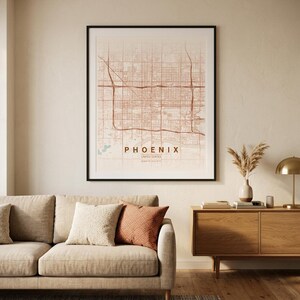 Puede incluir: Un mapa enmarcado de Phoenix, Arizona, en una paleta de colores neutros, cuelga sobre un sofá beige con cojines decorativos. El mapa está en un marco negro y presenta el diseño de las calles de la ciudad. También se ven un mueble de madera y una lámpara.