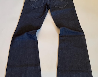 May include: Dark wash denim jeans with a flared leg design. The jeans feature classic five-pocket styling, a button and zipper closure, and a leather patch on the back. The jeans are laid flat on a light surface.