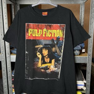 May include: Black t-shirt featuring a graphic from the movie "Pulp Fiction." The graphic shows a woman with a cigarette, and the movie title is in yellow and red. The shirt is hanging on a metal rack.