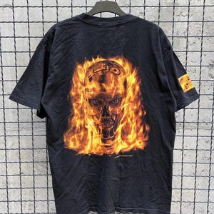 Könnte beinhalten: Schwarzes T-Shirt mit einem flammenden Schädel-Motiv. Das Design zeigt einen von Flammen umgebenen Schädel, mit dem Text "UNIVERSAL STUDIOS" auf dem Ärmel. Das T-Shirt hängt an einem schwarzen Kleiderbügel vor einem Drahtgitterhintergrund.