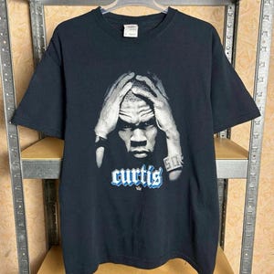 Könnte beinhalten: Schwarzes T-Shirt mit einem Graustufenbild einer Person mit den Händen am Kopf. Das Wort "curtis" ist in Blau und Weiß gedruckt, darunter eine kleine Krone. Die Zahl "50" ist am Handgelenk zu sehen.