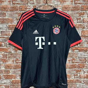 May include: Black athletic jersey with red accents, featuring the Adidas logo and a team emblem. The jersey has red stripes on the shoulders and red trim on the sleeves. The team's logo is on the chest.
