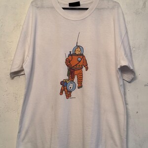 以下が含まれることがあります： オレンジ色の宇宙服を着た人物と、お揃いの宇宙服を着た犬の漫画イラストが描かれた白いTシャツ。イラストには青と黄色のアクセントがあります。Tシャツは黒いハンガーに吊るされています。