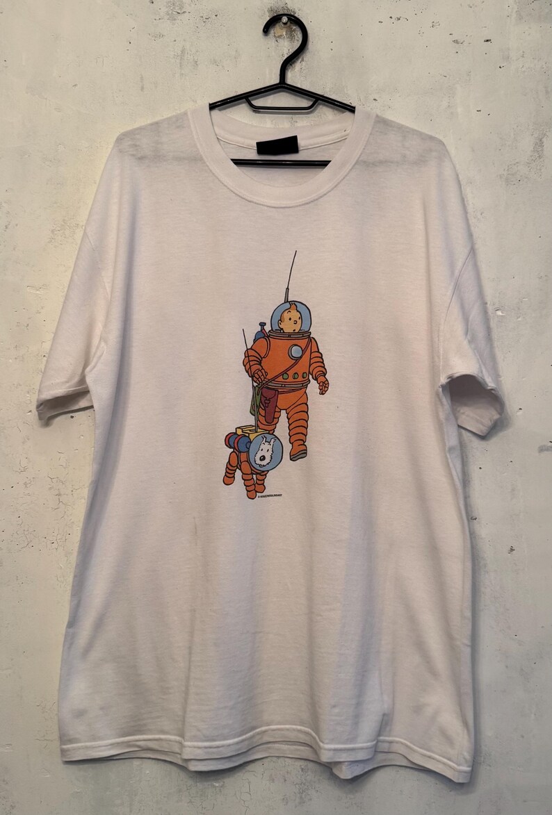 以下が含まれることがあります： オレンジ色の宇宙服を着た人物と、お揃いの宇宙服を着た犬の漫画イラストが描かれた白いTシャツ。イラストには青と黄色のアクセントがあります。Tシャツは黒いハンガーに吊るされています。
