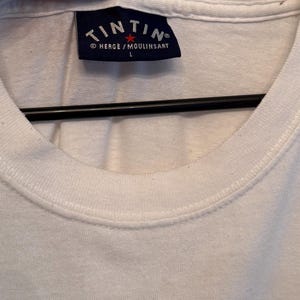 以下が含まれることがあります： 白のクルーネックTシャツ。紺色の長方形のラベルが付いています。ラベルには、白いアーチ状のテキストで「TINTIN」の文字が、その下に「HERGE / MOULINSART」と文字「L」が記載されています。
