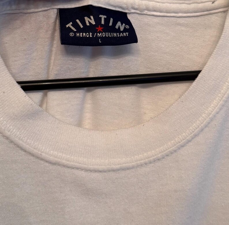 以下が含まれることがあります： 白のクルーネックTシャツ。紺色の長方形のラベルが付いています。ラベルには、白いアーチ状のテキストで「TINTIN」の文字が、その下に「HERGE / MOULINSART」と文字「L」が記載されています。