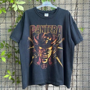 Puede incluir: Camiseta negra con el nombre de la banda "PANTERA" en letras doradas y rojas. El gráfico representa una figura demoníaca con cuernos y una calavera. La camiseta está colgada en una percha sobre un fondo de malla metálica.