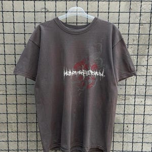 May include: A dark gray t-shirt with the text "HEAVEN SHALL BURN" in white, gothic-style lettering, and a red floral design. The shirt is hanging on a hanger against a wire mesh background.