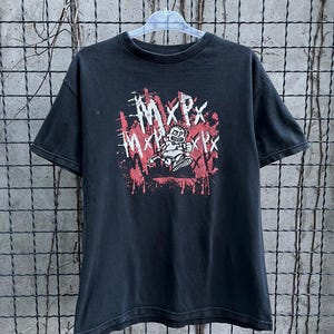 Peut inclure: T-shirt noir avec le nom du groupe "MxPx" dans un design vieilli de style graffiti avec des accents rouges. Le graphisme comprend un personnage de dessin animé et est imprimé sur un tee-shirt à manches courtes.
