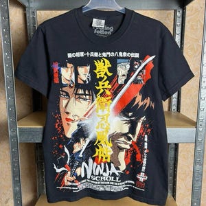 May include: Black t-shirt featuring a vibrant graphic from "Ninja Scroll." The design includes Japanese text and illustrations of characters with swords and action scenes. The shirt is hanging on a hanger.