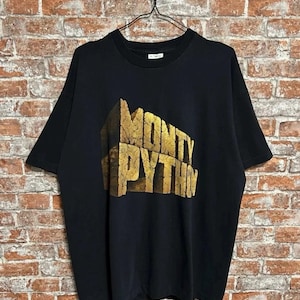 Puede incluir: Camiseta negra con el texto dorado "MONTY PYTHON" en letras de bloque 3D. La camiseta está colgada de una percha negra sobre un fondo de pared de ladrillo. La camiseta es de estilo cuello redondo clásico.