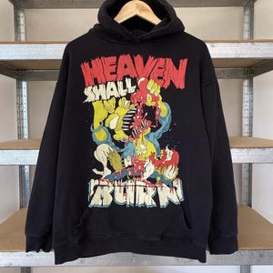 Puede incluir: Sudadera con capucha negra con un estampado gráfico de un personaje de dibujos animados con el texto "Heaven Shall Burn".