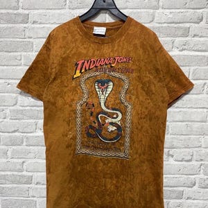 Pode incluir: Camiseta marrom com o texto "Indiana Jones Adventure" e um gráfico de cobra dentro de uma moldura decorativa. A camiseta está pendurada em um cabide preto contra uma parede de tijolos brancos. A cor da camiseta é quente.