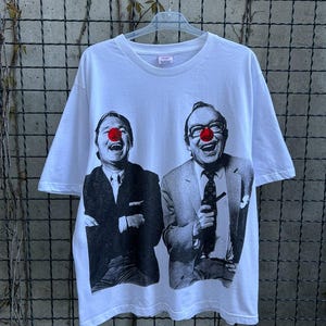 Peut inclure: T-shirt blanc avec une image en noir et blanc de deux hommes en costume, portant chacun un nez de clown rouge et riant. Le t-shirt a des manches courtes et un col rond.