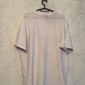 以下が含まれることがあります： 黒いハンガーに掛けられた無地の白いTシャツ。背景は、テクスチャ加工されたオフホワイトの壁です。Tシャツは柔らかい素材でできており、半袖です。このシャツは、ベーシックでカジュアルな衣服です。