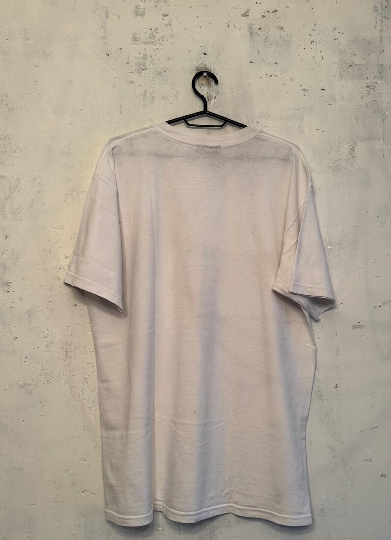以下が含まれることがあります： 黒いハンガーに掛けられた無地の白いTシャツ。背景は、テクスチャ加工されたオフホワイトの壁です。Tシャツは柔らかい素材でできており、半袖です。このシャツは、ベーシックでカジュアルな衣服です。