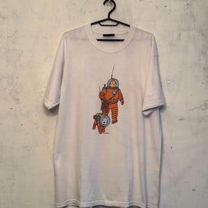 以下が含まれることがあります： 宇宙飛行士と宇宙服を着た犬の漫画イラストが描かれた白いTシャツ。宇宙飛行士はオレンジ色で、犬はオレンジと青色です。Tシャツは、ニュートラルな壁を背景に、黒いハンガーに掛けられています。