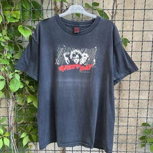 May include: Black t-shirt featuring a band graphic with the text "Green Day" in red. The shirt has a crew neck and short sleeves. The band graphic is in white and black. The shirt is hanging on a hanger.