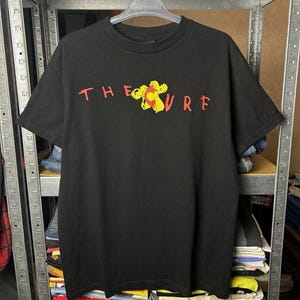 May include: Black t-shirt with the text "THE CURE" in red, with a yellow and red floral graphic. The shirt is hanging on a metal rack.