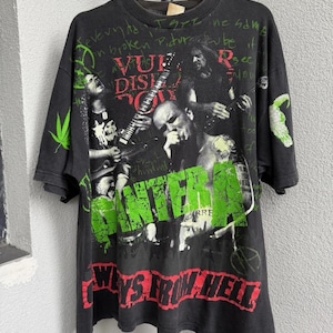 Può includere: T-shirt nero vintage con grafica di una band. Il design presenta il nome della band in verde, con le parole "Ways From Hell" in rosso. La maglietta ha un aspetto invecchiato e include immagini dei membri della band e altre grafiche.