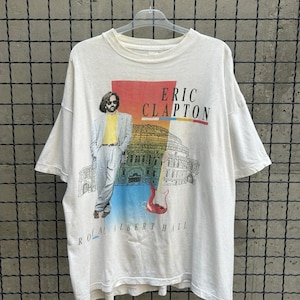 Puede incluir: Camiseta blanca con un gráfico de Eric Clapton, el texto "Eric Clapton" en rojo y "Royal Albert Hall" debajo de una ilustración de un edificio y una guitarra. La camiseta tiene mangas cortas y cuello redondo.