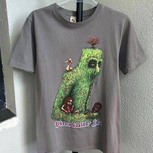May include: Gray t-shirt featuring a colorful graphic of a green, moss-covered creature with a tree on its head, a duck, and two figures. The text "Dinosaur Jr" is printed below the graphic in a colorful font.