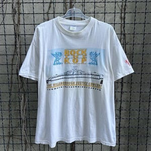 May include: White t-shirt featuring the text "ROCK THE KOP" above a graphic of a bridge and the text "THE HILLSBOROUGH JUSTICE CONCERT TENMAYNINETEENINETYSEVEN". The shirt has short sleeves and a crew neck.