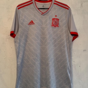 May include: A light gray short-sleeved soccer jersey featuring a subtle geometric pattern, red Adidas logo on the right chest, and the Spain national team crest with a red star on the left chest. The V-neck collar and sleeve cuffs are red.