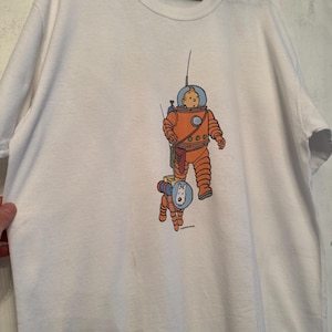 以下が含まれることがあります： 宇宙飛行士と犬の漫画イラストが描かれた白いTシャツ。宇宙飛行士はオレンジ色の宇宙服と青いヘルメットを着用しています。犬もオレンジ色の宇宙服と青いヘルメットを着用しています。