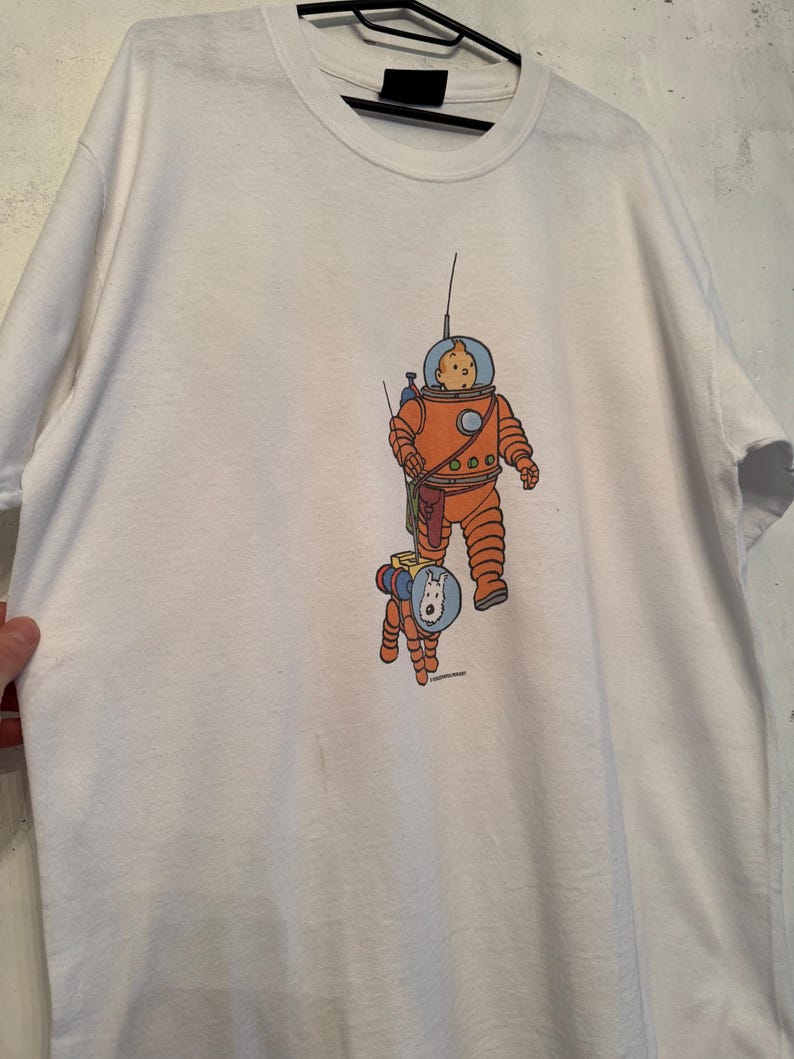 以下が含まれることがあります： 宇宙飛行士と犬の漫画イラストが描かれた白いTシャツ。宇宙飛行士はオレンジ色の宇宙服と青いヘルメットを着用しています。犬もオレンジ色の宇宙服と青いヘルメットを着用しています。