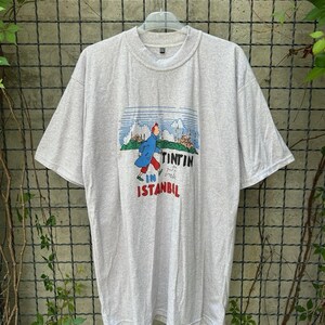 May include: A light gray t-shirt featuring a graphic of Tintin in Istanbul. The graphic shows Tintin in a blue coat, walking with a dog, with the words "Tintin in Istanbul" in red and black. The shirt is hanging on a metal grid.