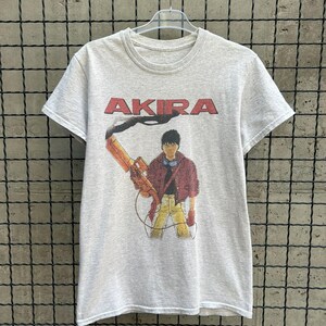 以下が含まれることがあります： 赤いブロック体の「AKIRA」の文字が入ったライトグレーのTシャツ。テキストの下には、武器を持っている人のグラフィックがあります。その人は赤いジャケット、黄色のズボン、手袋を着用しています。Tシャツは透明なプラスチックハンガーにかかっています。