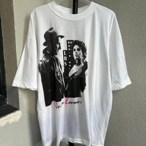 May include: White t-shirt featuring a black and white graphic of two people, one wearing a hat and coat, the other in a dress. The text "LBS" and "Victor Loomes" are also visible on the shirt.