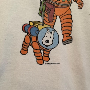 以下が含まれることがあります： オレンジ色の宇宙服を着たキャラクターと、お揃いの宇宙服を着た犬の漫画イラストが描かれた白いTシャツ。犬は青い円の中にいます。テキスト「HERGE/MOULINSART」が下部に印刷されています。