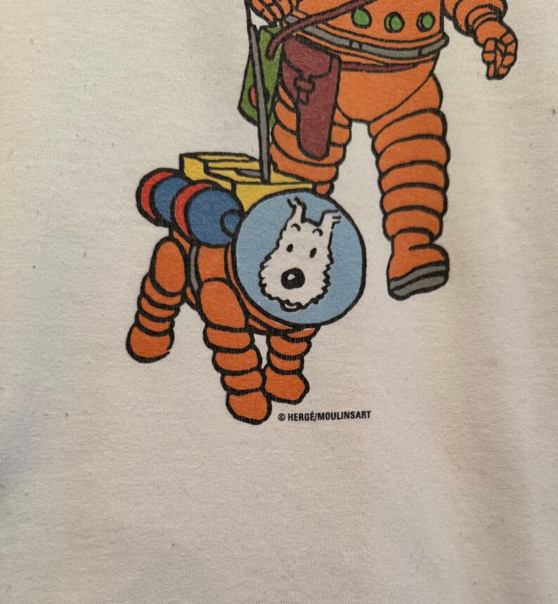 以下が含まれることがあります： オレンジ色の宇宙服を着たキャラクターと、お揃いの宇宙服を着た犬の漫画イラストが描かれた白いTシャツ。犬は青い円の中にいます。テキスト「HERGE/MOULINSART」が下部に印刷されています。