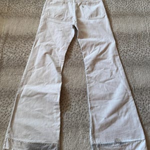 May include: Light gray flared jeans with two back pockets and a button closure. The pants have a high waist and a wide leg opening. The fabric appears to be a light-weight denim. The jeans are laid flat on a textured surface.