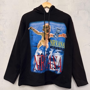 Puede incluir: Una sudadera negra con capucha y manga larga que presenta un estampado gráfico de la portada del álbum Nevermind de Nirvana. El diseño incluye un bebé nadando, un billete de dólar y el texto Nirvana y Nevermind en letras amarillas, rojas y negras.