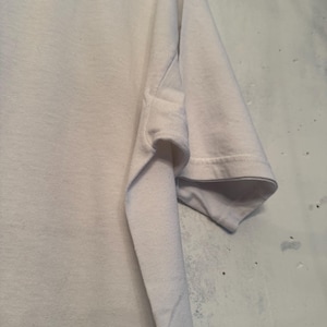 以下が含まれることがあります： 白いTシャツの袖のクローズアップ。 袖口で折りたたまれ、内側の生地が見えています。 生地は柔らかく、軽量な素材のようです。 背景は、質感のある明るい色の壁です。